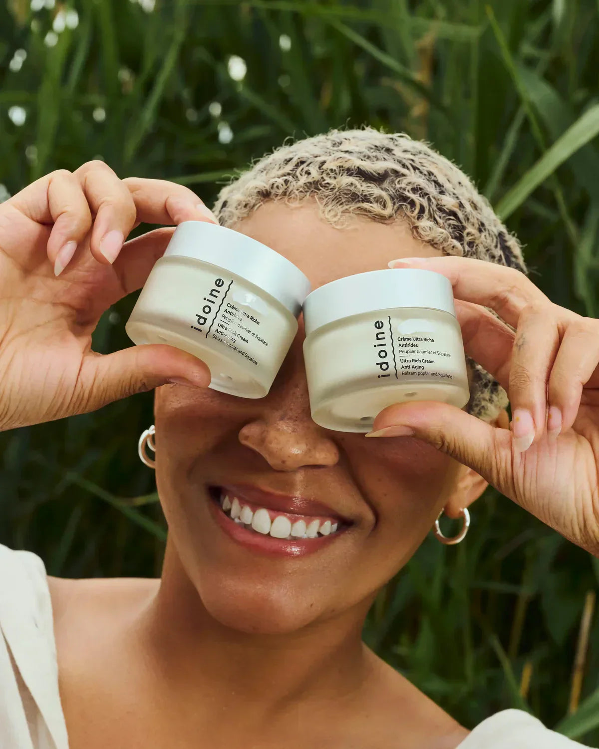 Crème antirides Idoine Bio Ultra Riche tenue devant les yeux par une femme souriante, fond végétal