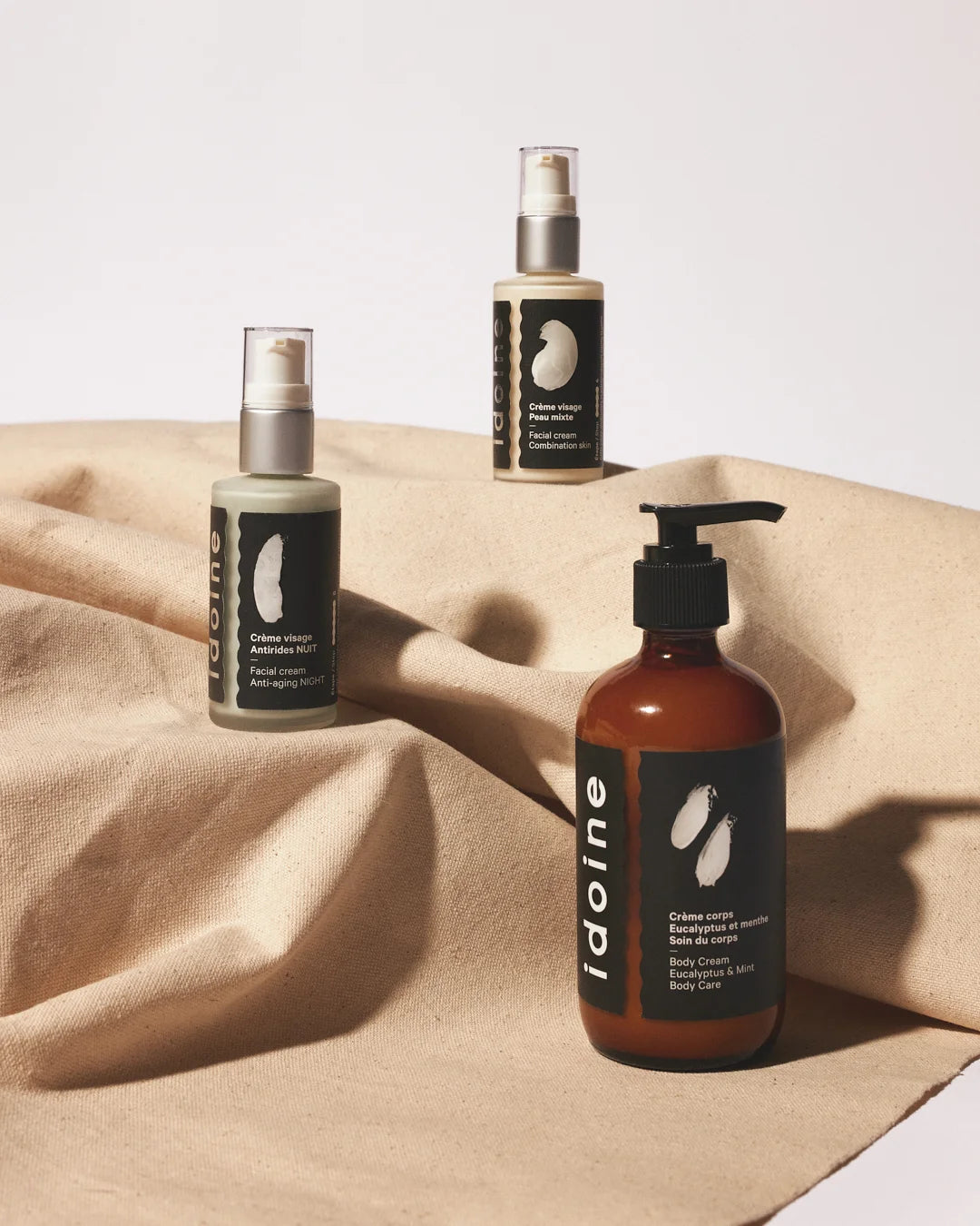 Idoine skincare products arranged on beige fabric, featuring two pump bottles of facial cream and one amber pump bottle of eucalyptus and mint body cream