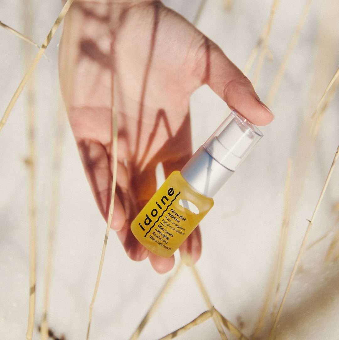 A hand holding a small yellow bottle of Idoine skincare serum with a pump dispenser, shown against a soft natural background with dried grass stems.