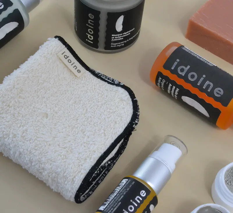 Flat lay of Idoine skincare products including a white washcloth with black trim, a soap bar, pump bottles, and jars with black labels, arranged on a beige surface.