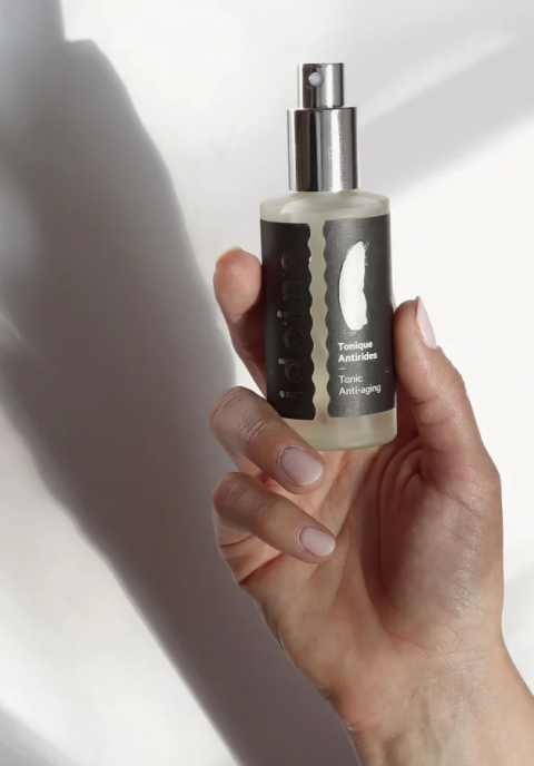 Spray tonique anti-âge pour le visage dans un flacon transparent avec pompe argentée, tenu à la main sur un fond de lumière douce.