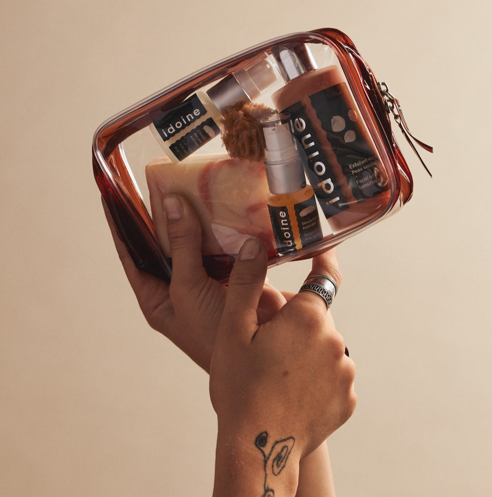 Trousse transparente contenant des produits Idoine Bio, tenue à deux mains, avec un tatouage visible sur le poignet.