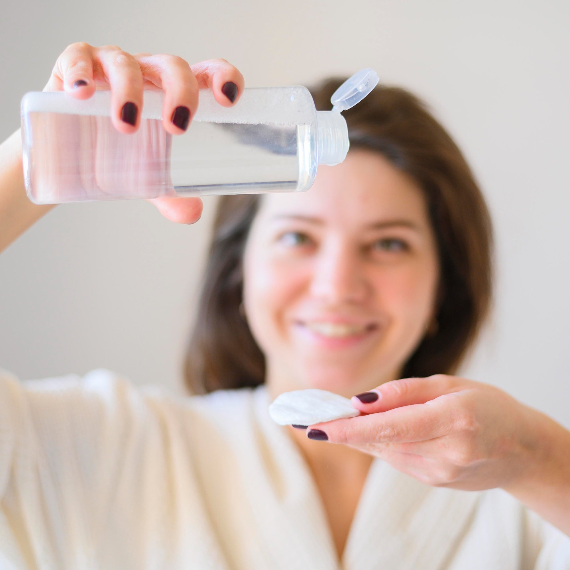 Front-facing portrait of a woman removing her makeup with a cotton pad, clean minimal backdrop