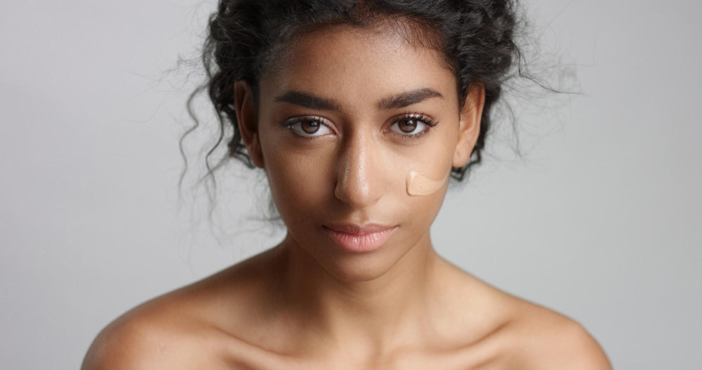 Portrait d’une femme à la peau mate appliquant du fond de teint sur sa joue, regard face caméra, fond neutre gris.