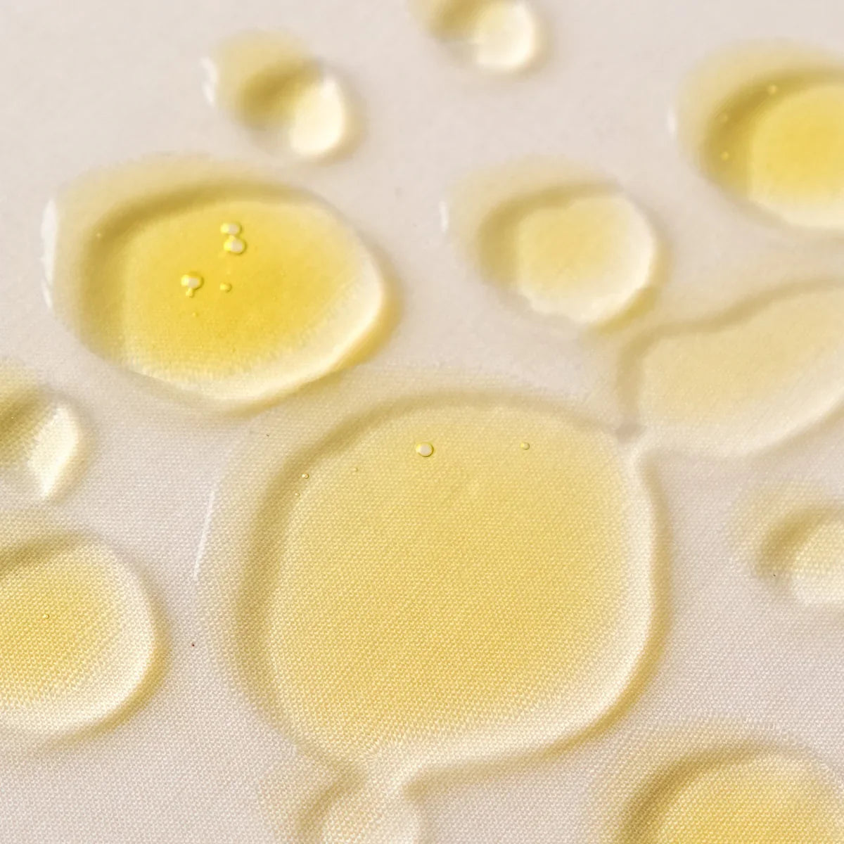 Gouttes d'huile visage jaune dorée étalées sur tissu beige, texture légère et fluide 
