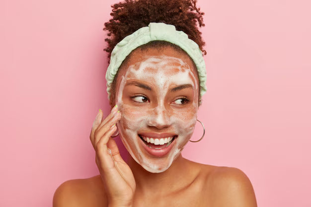 Femme souriante avec bandeau vert nettoie son visage avec une mousse nettoyante, fond rose, routine de soin du visage