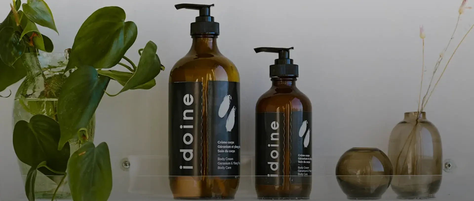 Idoine natural body cream bottles with pump dispensers on shelf beside plants and vases.