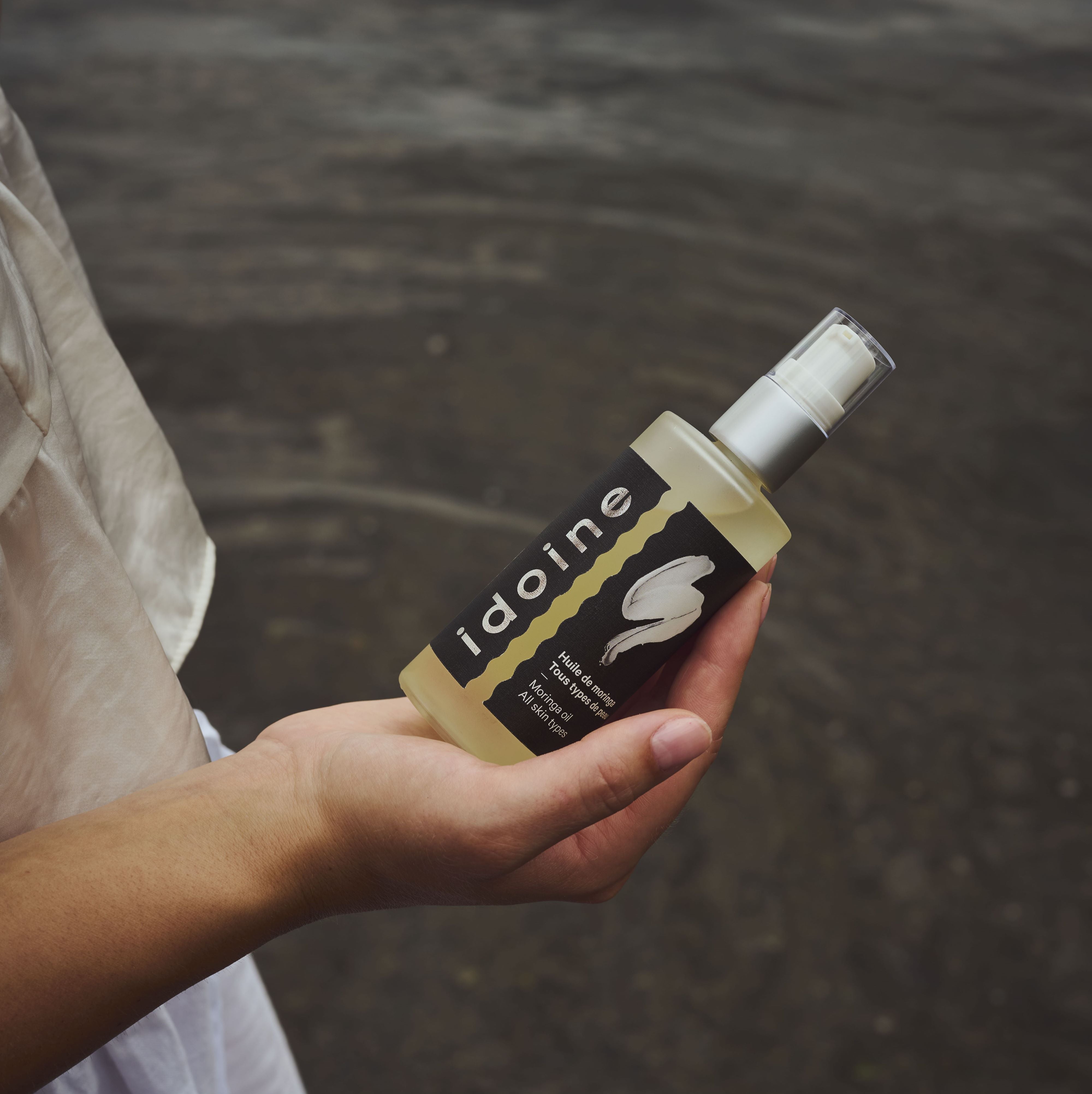 Idoine Bio moringa body oil in a glass bottle with black label and silver pump, held by a woman outdoors, designed to nourish and suit all skin types.